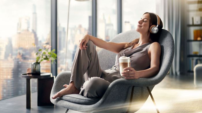 אישה יושבת על כורסה, מחייכת, עם אוזיות על ראשה וכוס קפה שהיא אוחזת בה