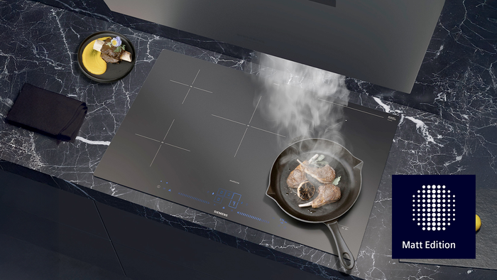         Elegantes Siemens Induktionskochfeld mit einer Pfanne mit gebratenem Lamm, umgeben von frischen Zitronen auf einer dunklen Marmoroberfl&auml;che.       