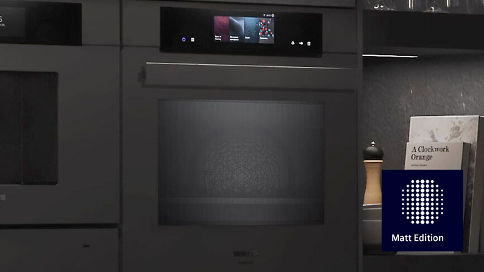         Ein eleganter schwarzer Siemens Ofen mit einem digitalen Display, begleitet von einer Pfefferm&uuml;hle, Sch&uuml;sseln und einem Buch mit dem Titel "Uhrwerk Orange".       