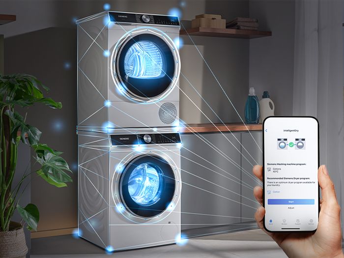 A hand holding a smartphone controlling a stacked Siemens washer and dryer, connected by glowing blue lines, in a modern laundry room.