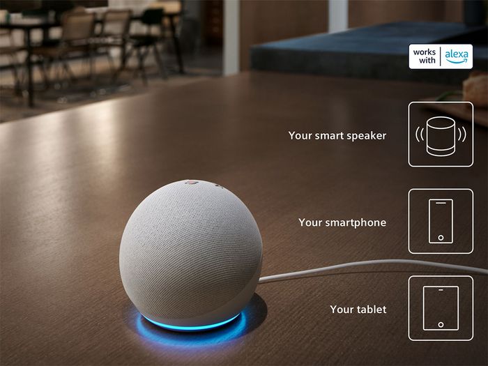 A smart speaker with a lit blue ring sits on a table. Icons for smartphone and tablet are displayed, along with a "works with Alexa" badge.