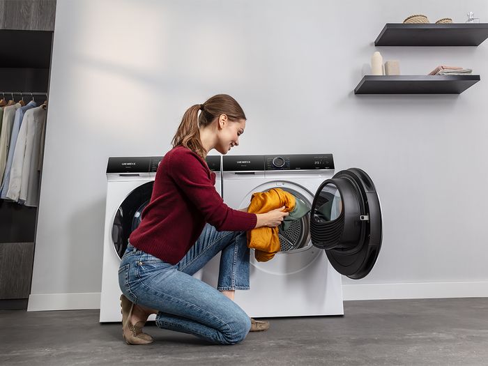 A woman kneels while loading colorful laundry into a modern dryer in a stylish, minimalistic laundry room.