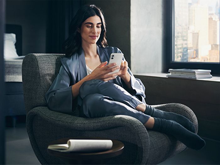 A woman sitting in an armchair, looking at her smartphone in a modern room with a city view.