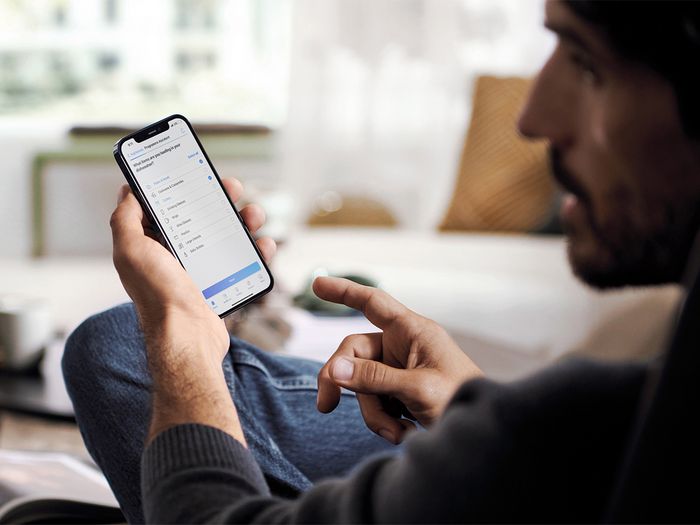 A man using a smartphone app to select a cooking program, with an open book and coffee table in the background.