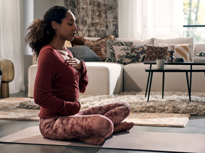 A woman meditating on a yoga mat in a quiet living room, with the Quiet Mark logo visible.