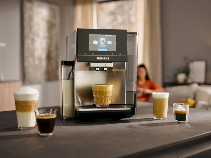 A Siemens coffee machine making a latte, surrounded by various coffee drinks and a plate of macarons, with a person relaxing in the background.
