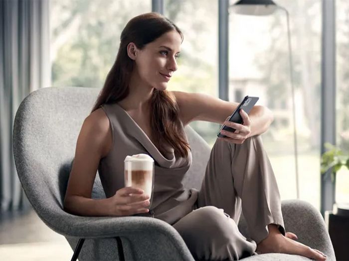 A woman relaxing in an armchair with a latte and a smartphone, looking out a large window.