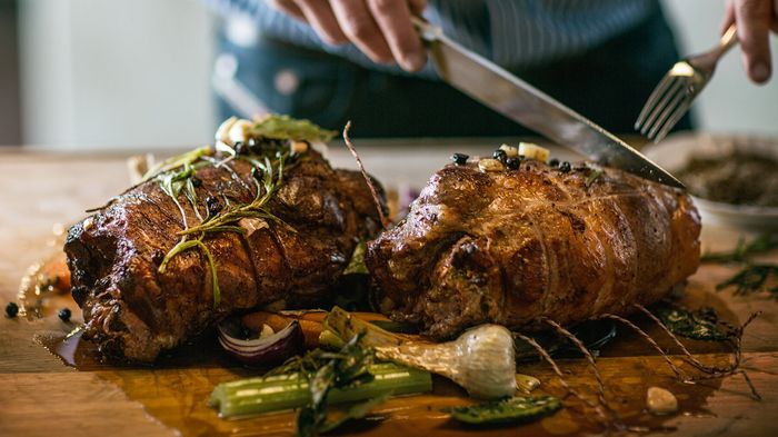 Persona che affetta, con coltello e forchetta su tagliere in legno, due arrosti di carne con rosmarino e verdure 