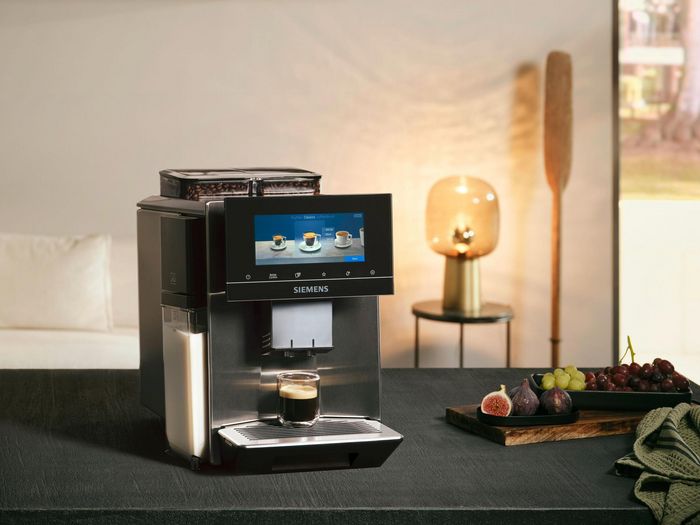 A Siemens coffee machine sits on a dark countertop in a stylish living room, with a cozy sofa and decorative items in view.