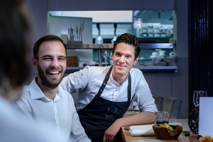 Uomo con grembiule che conversa con amici seduti al tavolo, con cucina a vista sullo sfondo