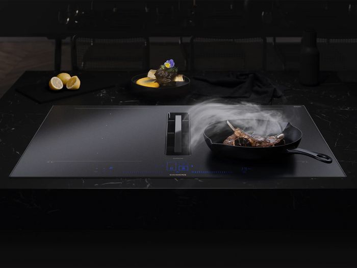 A black Siemens induction cooktop with an integrated downdraft extractor actively pulling steam from a cast iron pan cooking lamb chops, set in a dark, modern kitchen.