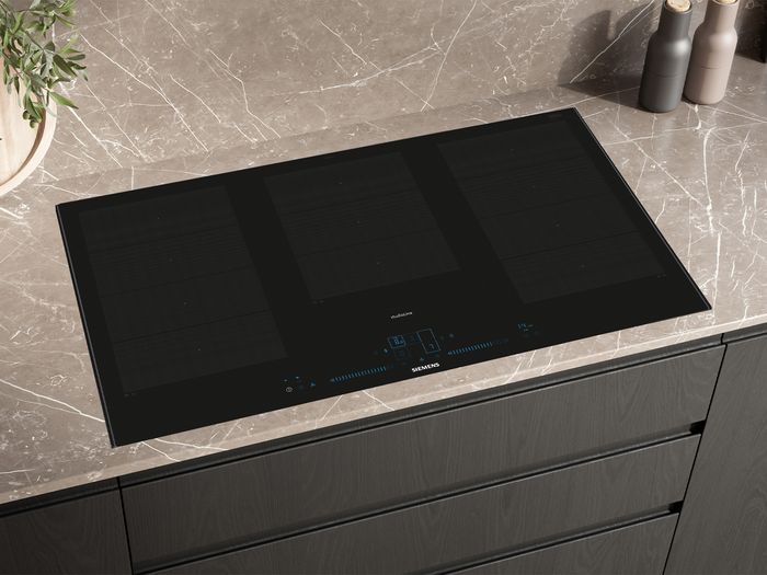 A black Siemens studioLine induction cooktop with multiple flexible cooking zones and blue digital controls, integrated into a dark marble-patterned countertop.