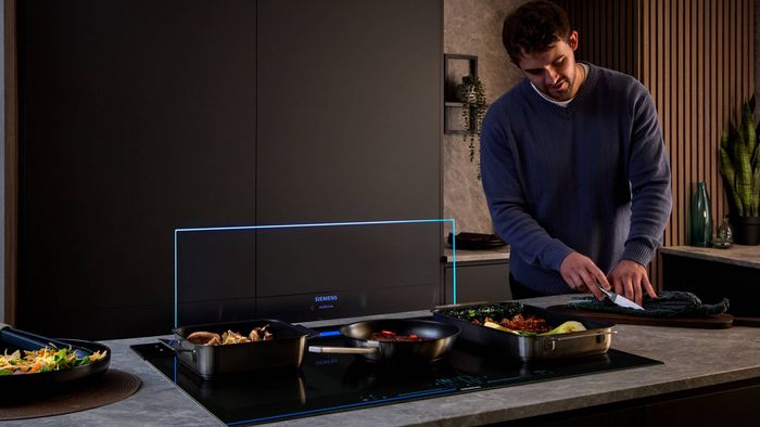 A man chops kale in a modern kitchen while cooking multiple dishes on a Siemens induction cooktop with an illuminated downdraft extractor.