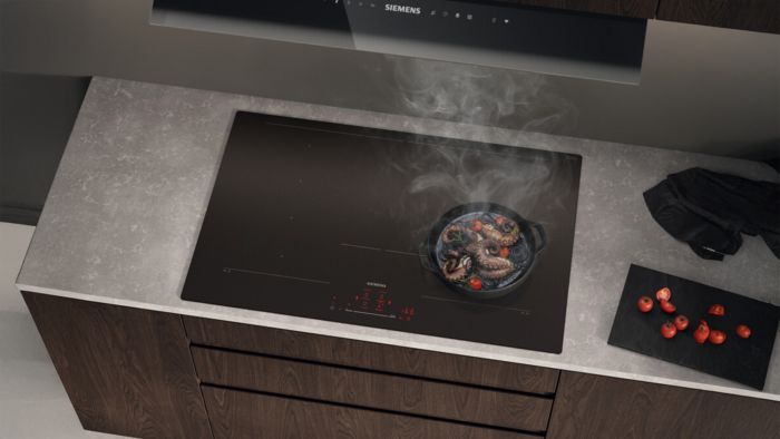 
       Une cuisine sombre avec une table de cuisson &agrave; induction noire, une po&ecirc;le de nourriture et une planche &agrave; d&eacute;couper avec des tomates.
      