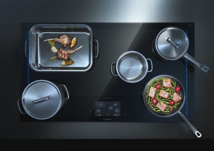 Vista dall&rsquo;alto di un piano cottura a induzione Siemens con pentole e padelle in cottura, contenenti carne, verdure e pesce