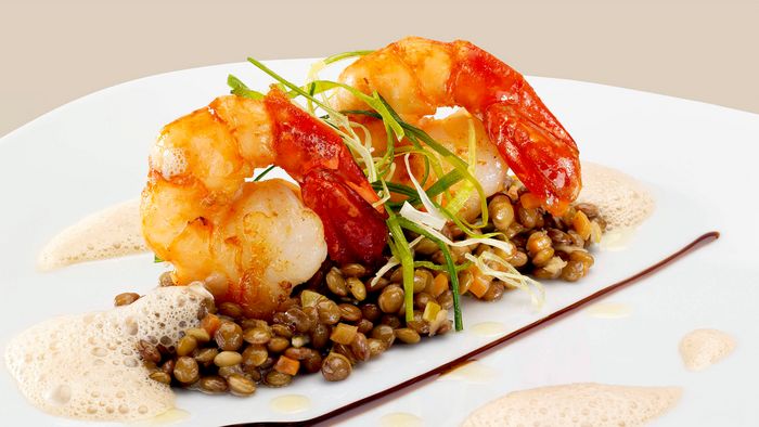 Een bord met een bedje van groenten zoals linzen, schuim eromheen en gegrilde garnalen bovenop.