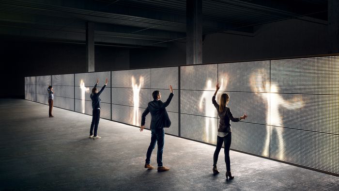 Tres figuras siluetadas con brazos levantados, de pie contra una pared oscura con efectos de iluminación.