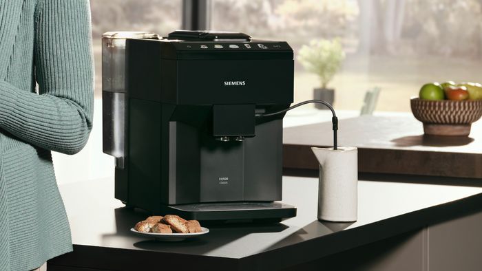 Siemens aparat za kafu na kuhinjskom pultu sipa mleko u šoljicu, a u blizini je tanjir s kolačićima